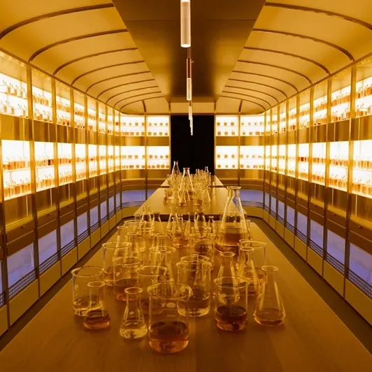 A long lab table filled with various glass beakers and flasks reflecting warm yellow lighting. Shelves of illuminated bottles line both walls, creating a symmetrical and glowing ambiance in the room.