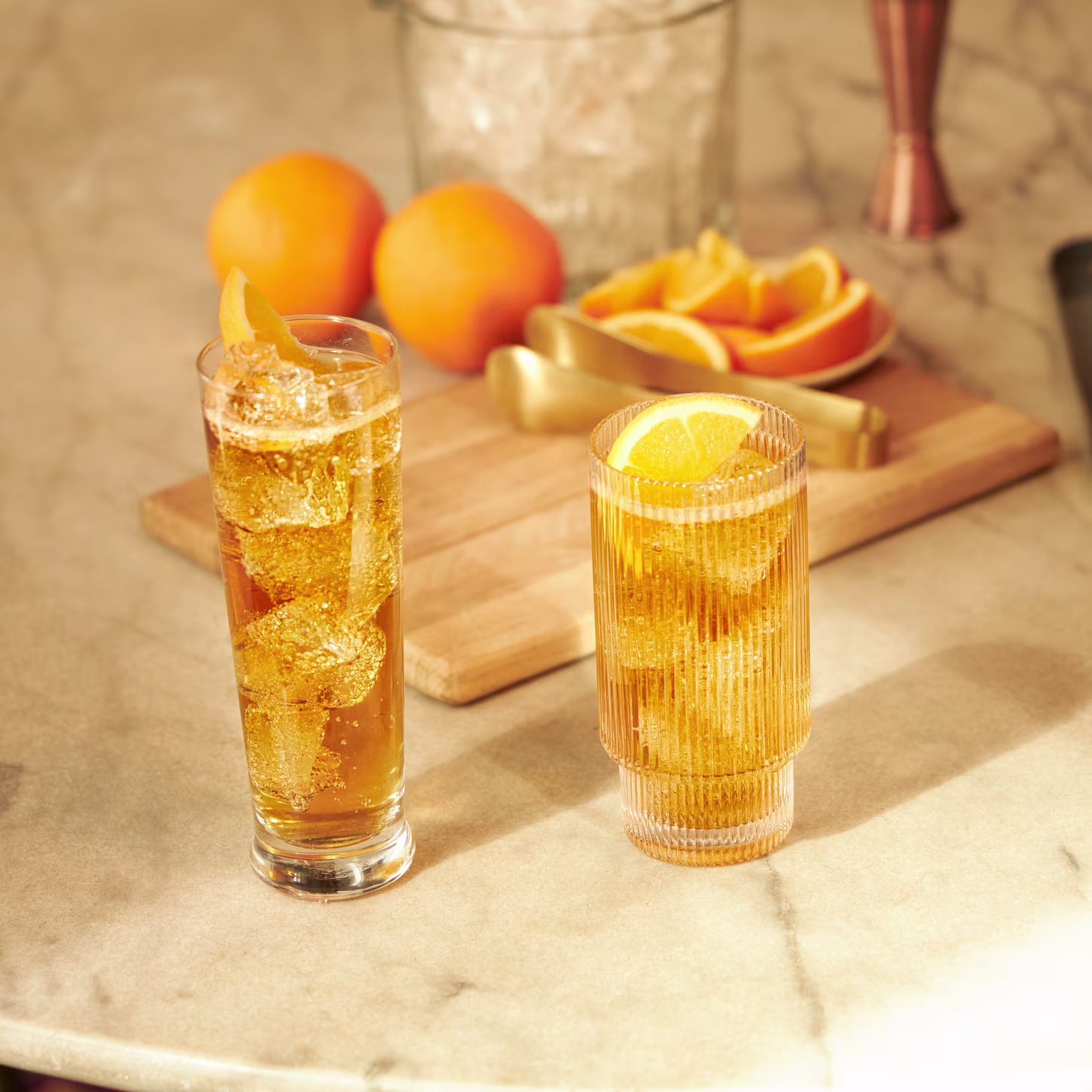 Two tall glasses filled with an orange-hued drink garnished with orange slices. A cutting board with sliced oranges, whole oranges, tongs, and an ice bucket are in the blurred background, all on a marble surface.