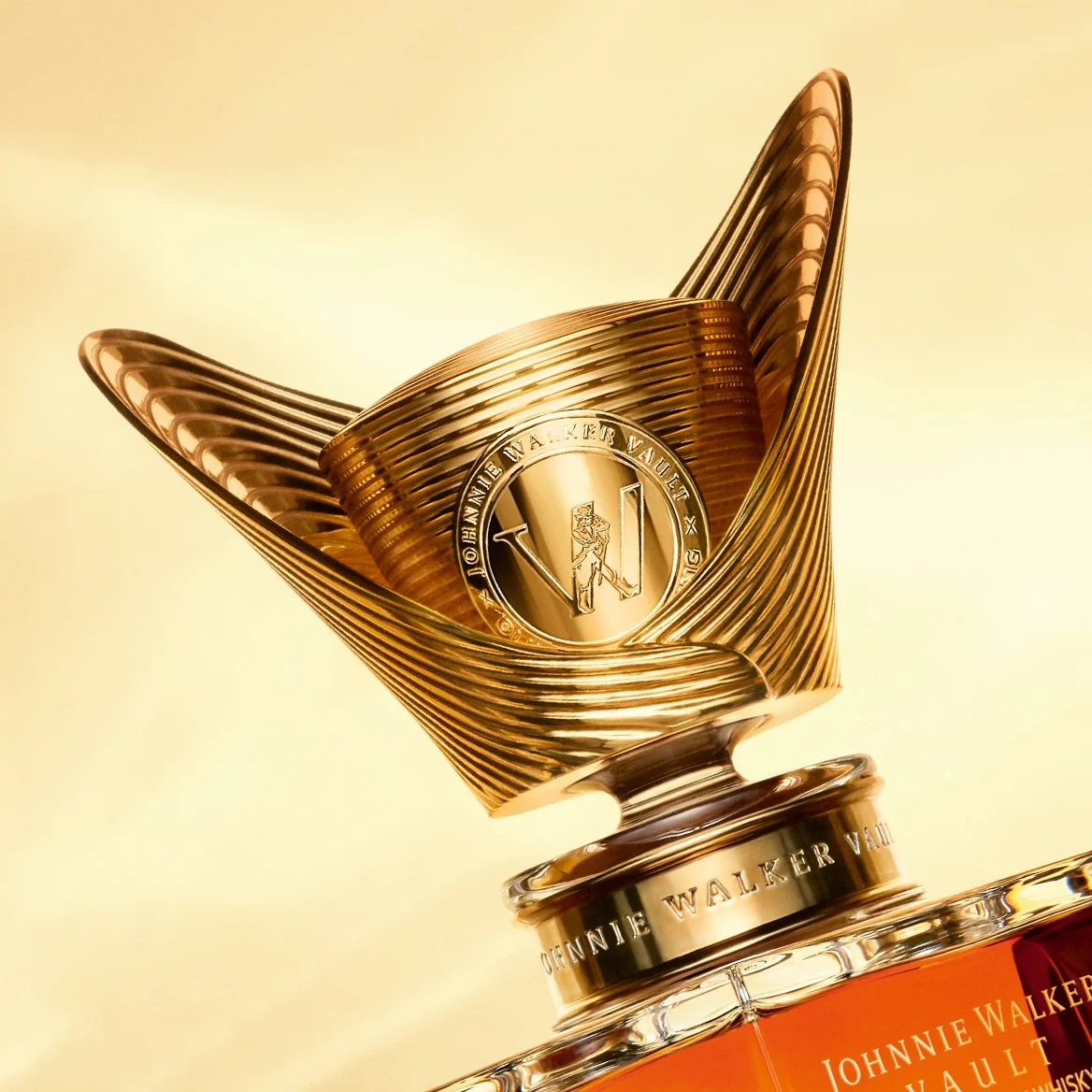 Close-up of a luxurious Johnnie Walker whisky bottle cap, featuring an ornate golden design with spiral elements and the Johnnie Walker logo embossed in the center, set against a warm, golden background.