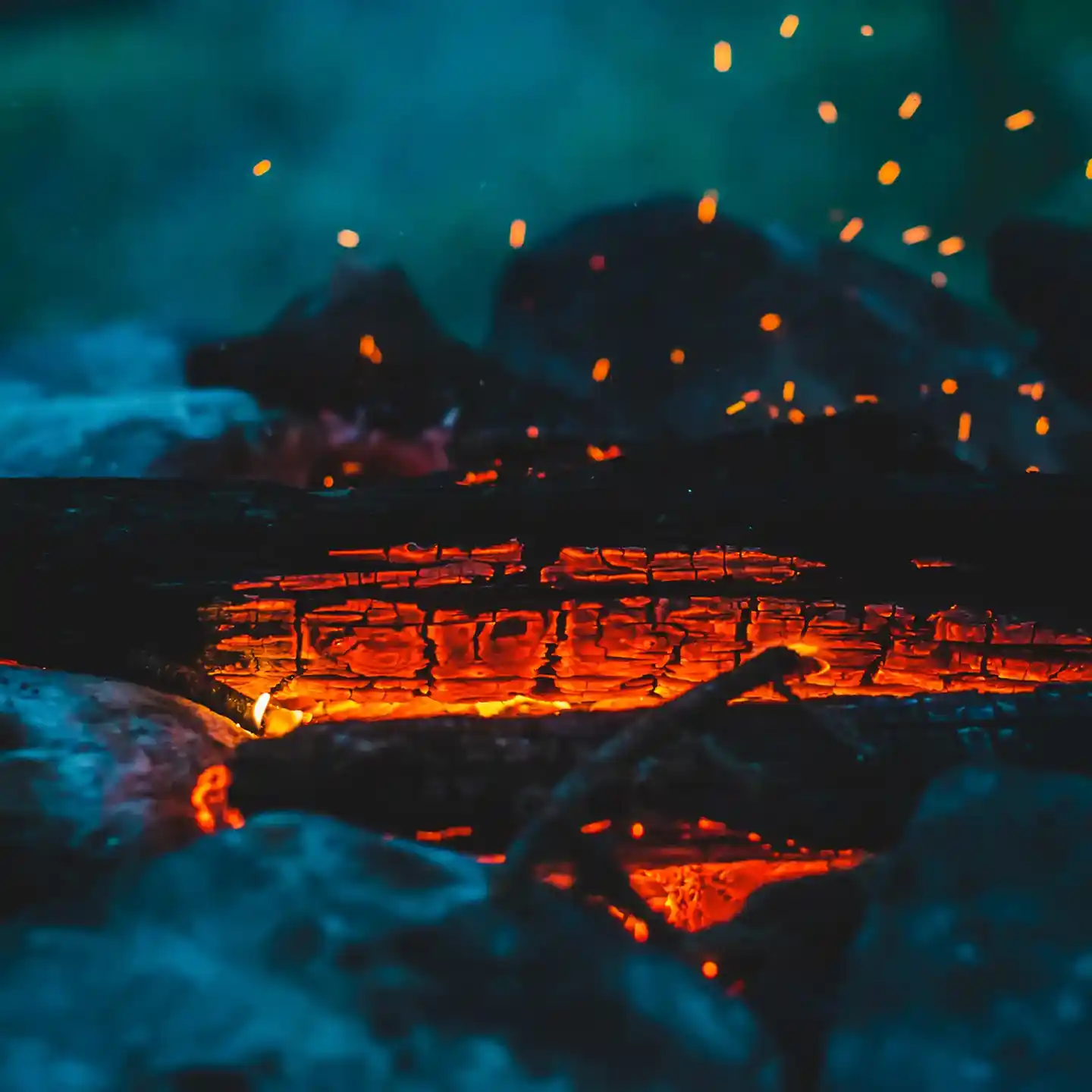 Close-up of a bonfire with glowing embers and charred wood. Bright orange sparks rise against a dark background, creating a warm, atmospheric scene.