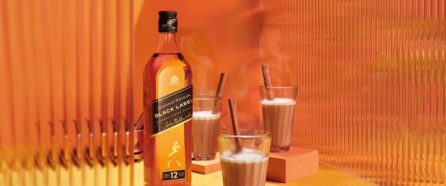 A bottle of Black Label whiskey sits on an orange textured surface. Beside her are three cups filled with steaming liquid, each with a cinnamon stick. The background features orange vertical lines, highlighting the warm and inviting atmosphere.