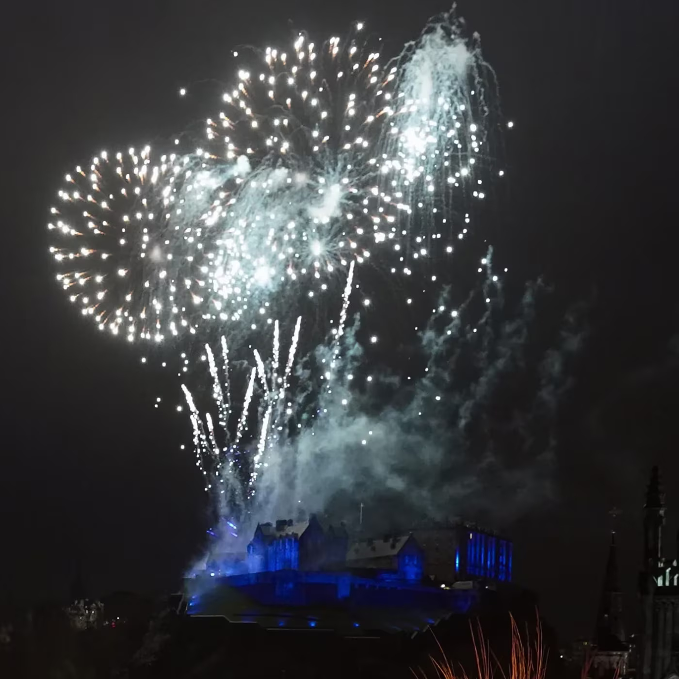 Fireworks burst in the night sky above a brightly lit castle with blue lighting.