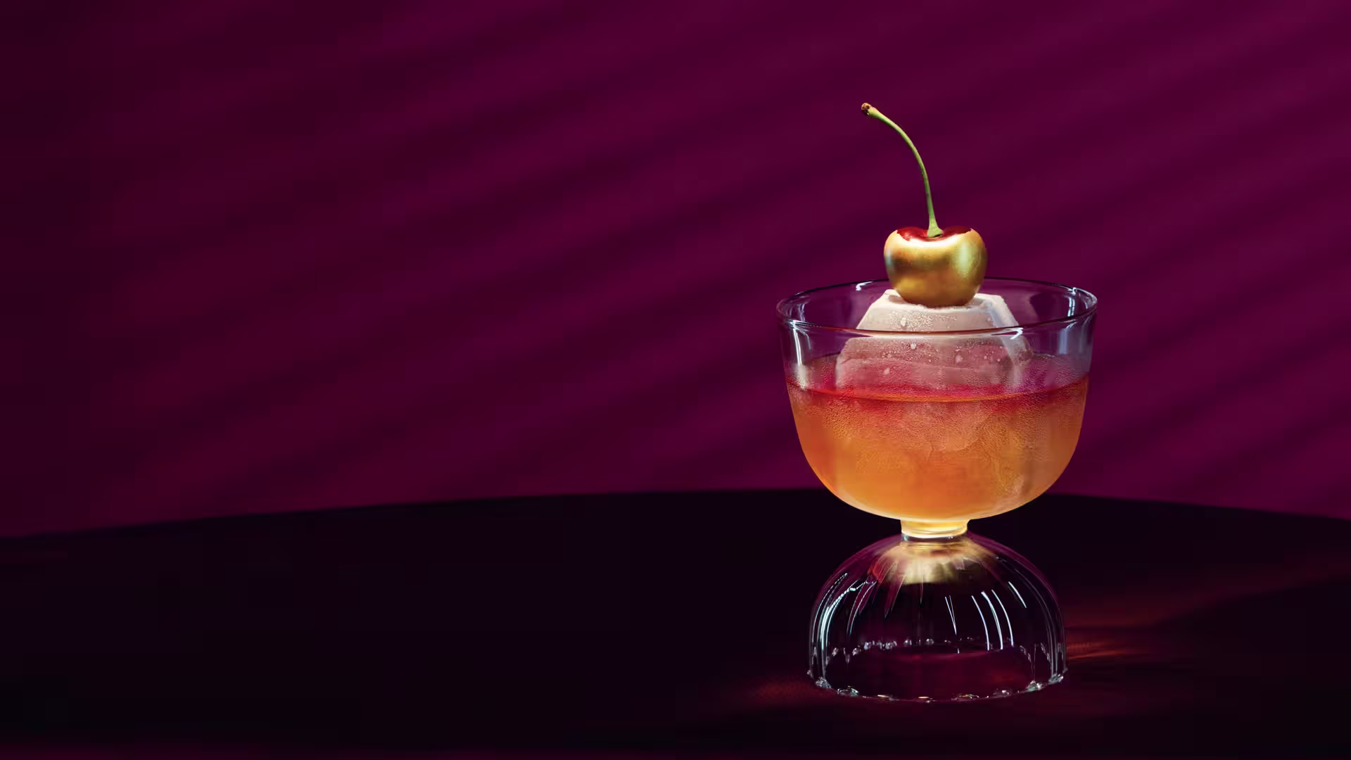 A cocktail in a glass with a large ice sphere and a cherry garnish sits on a table against a deep burgundy background with diagonal shadow lines.