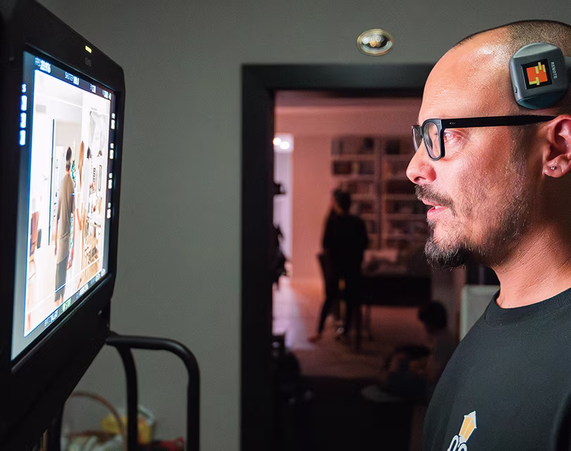 Un hombre con gafas y auriculares mira fijamente a un monitor que muestra imágenes, posiblemente de un plató de cine. La habitación está poco iluminada y al fondo hay una puerta en la que se ve la silueta de una persona.