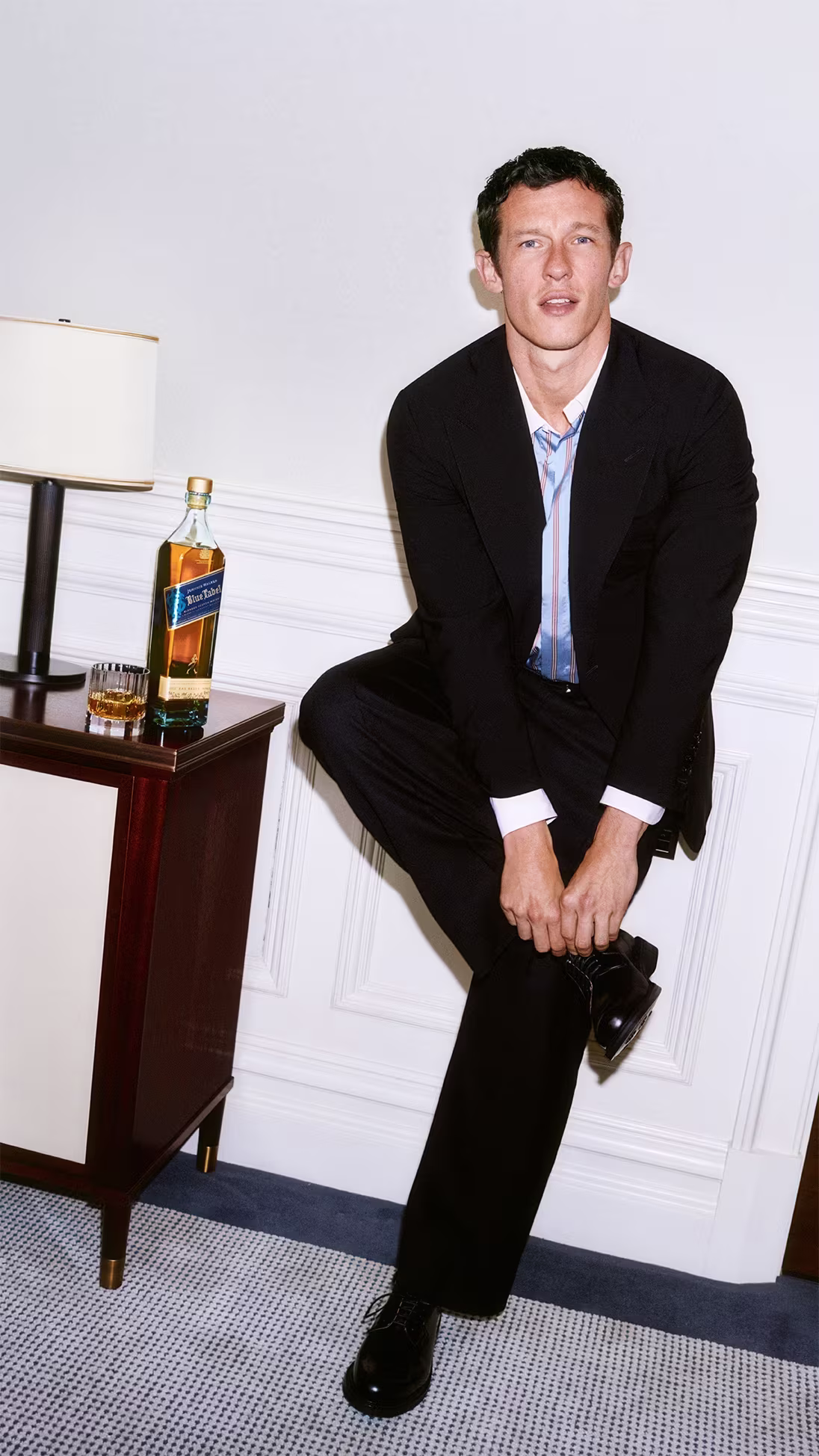 Callum Turner in a suit adjusts his shoe next to a table with a whiskey bottle, glass, and a lamp.