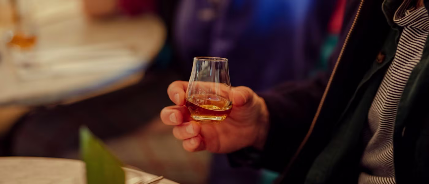 A person holds a small glass of whiskey. In the background you can see a blurry, set table with a plant and another person in a blue shirt. The atmosphere seems relaxed and sociable.