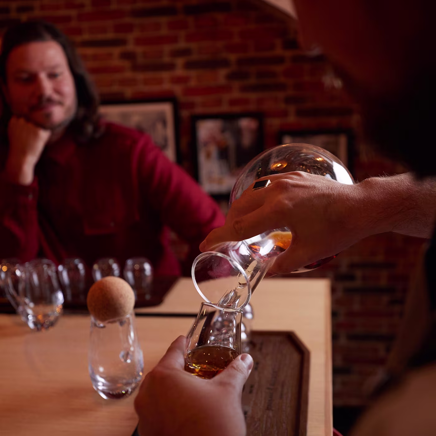 A person pours whiskey into a glass as another person watches at a table with brick walls in the background.