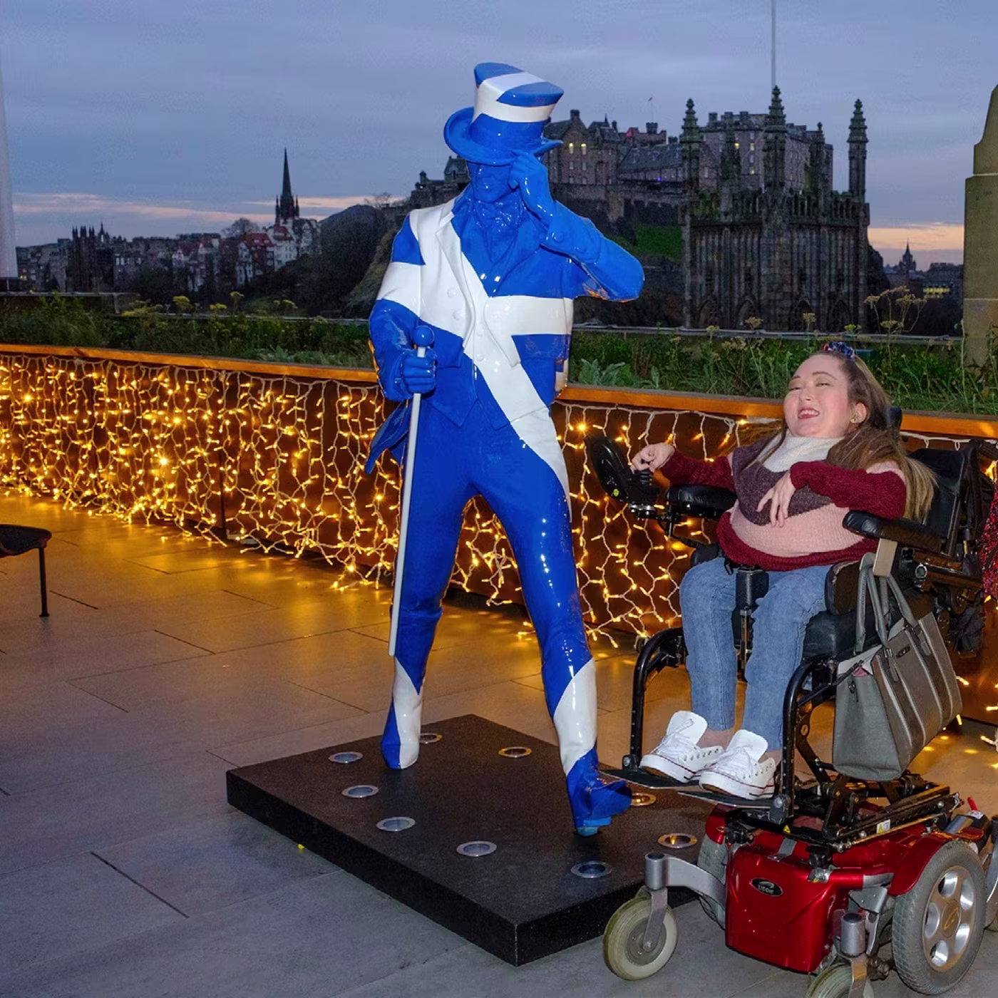 A smiling girl in a wheelchair poses with a blue-and-white statue in front of festive string lights outside.