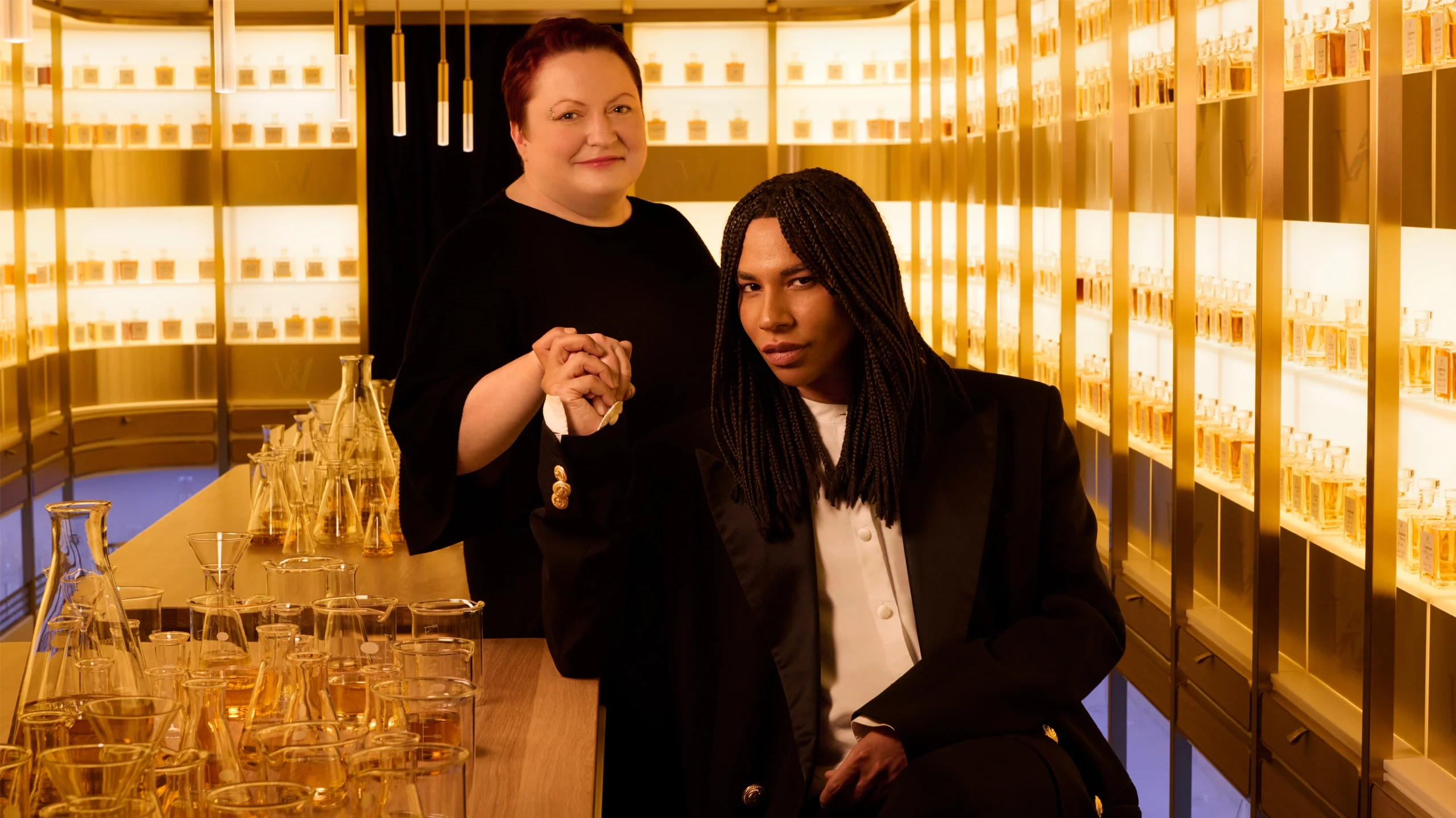 Two people pose in a luxurious, golden-lit room filled with glass perfume bottles and beakers. One stands smiling behind the other, who is seated, both holding hands and looking at the camera.