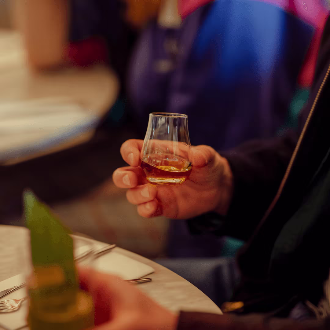 A person holds a small glass of whiskey. In the background you can see a blurry, set table with a plant and another person in a blue shirt. The atmosphere seems relaxed and sociable.