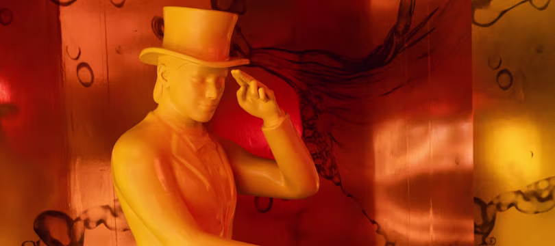 Una estatua amarilla de una persona que viste un sombrero de copa y un traje inclina su sombrero, sobre un fondo cálido de color naranja y rojo con patrones abstractos y giratorios.
