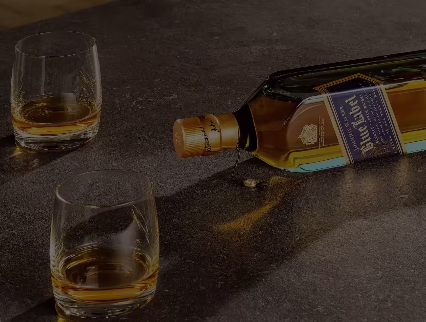 A bottle of Johnnie Walker whisky lying on its side, spilling some liquid, with two glasses of whisky on a dark stone surface.