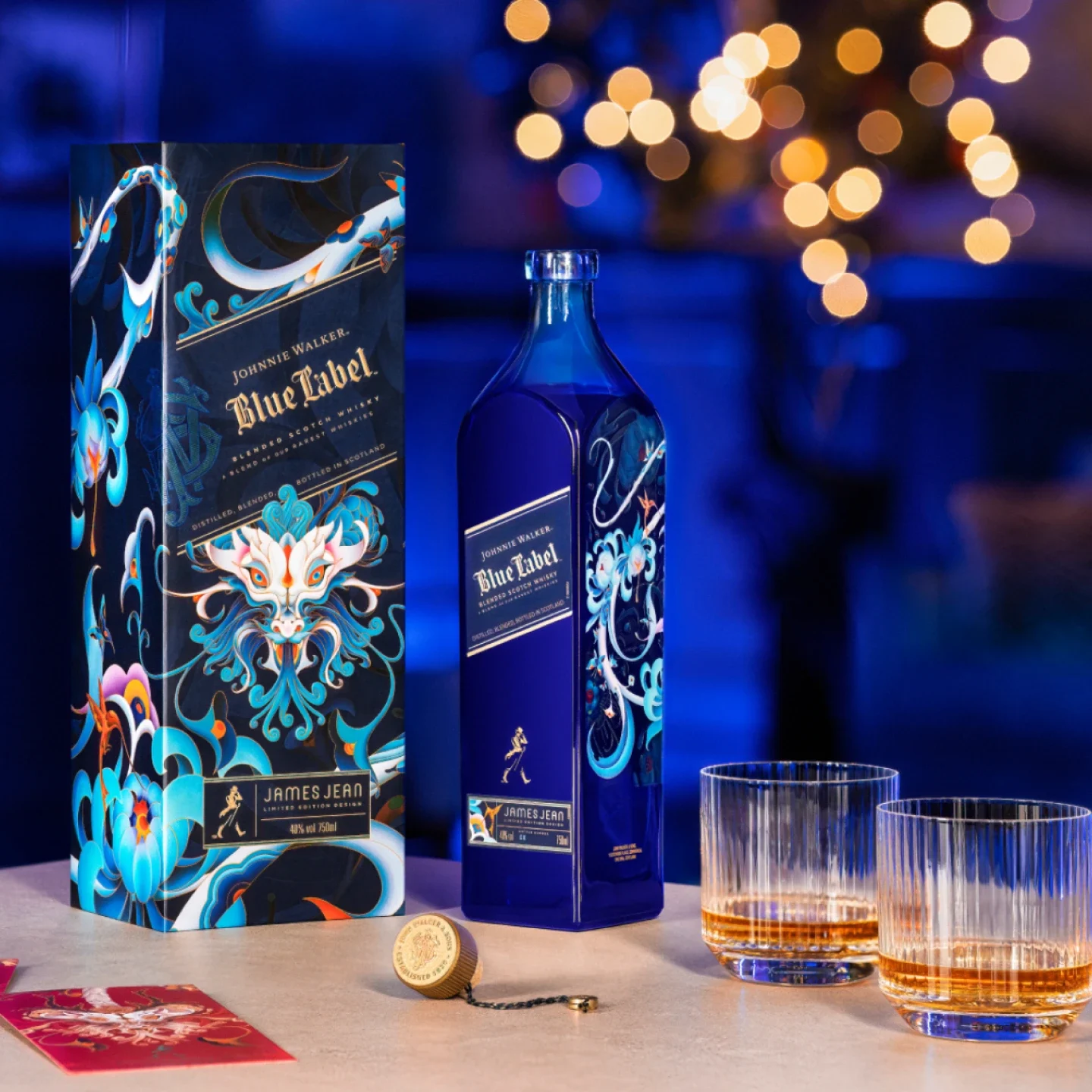Une bouteille bleue de whisky Johnnie Walker Blue Label avec une boîte à fleurs ornée est posée sur une table à côté de deux verres de whisky, d'un bouchon de bouteille et de lumières chaudes floues en arrière-plan.