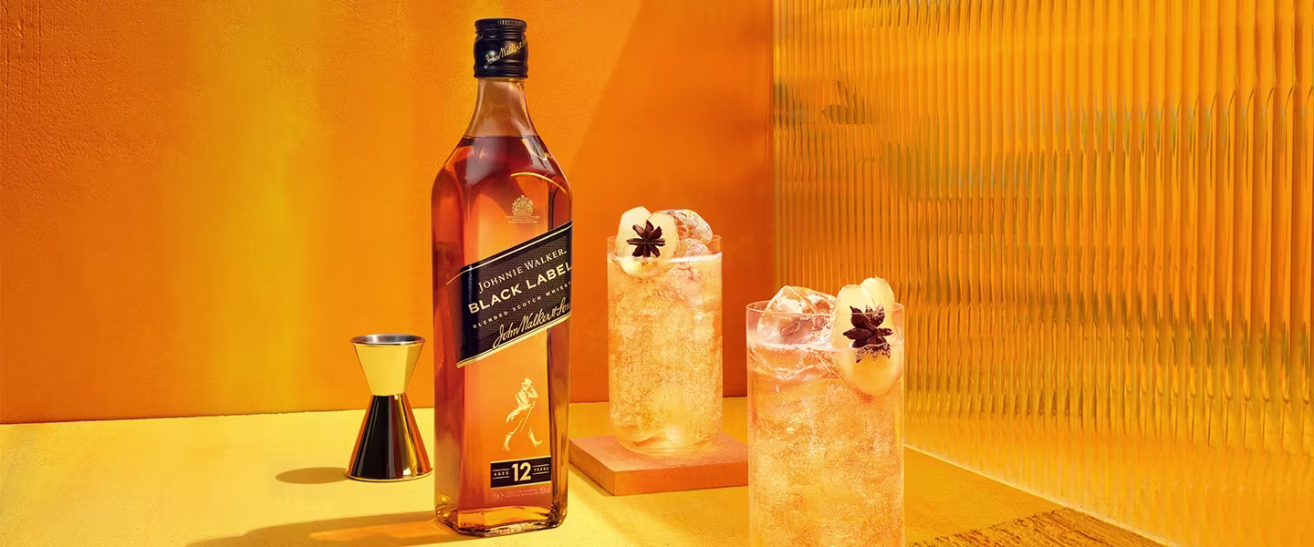  Eine Flasche Johnnie Walker Black Label mit zwei Cocktails auf Eis, garniert mit Sternanis, vor gelbem Hintergrund.