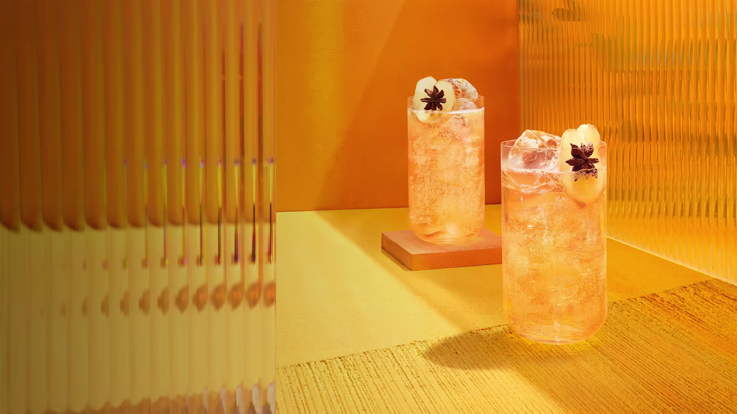 Zwei hohe Gläser mit einem hellorangen, eisgefüllten Cocktail, garniert mit Litschifrüchten und Sternanis, stehen auf einer strukturierten gelb-orangen Oberfläche mit warmer, moderner Beleuchtung und gewellten Glasplatten im Hintergrund.