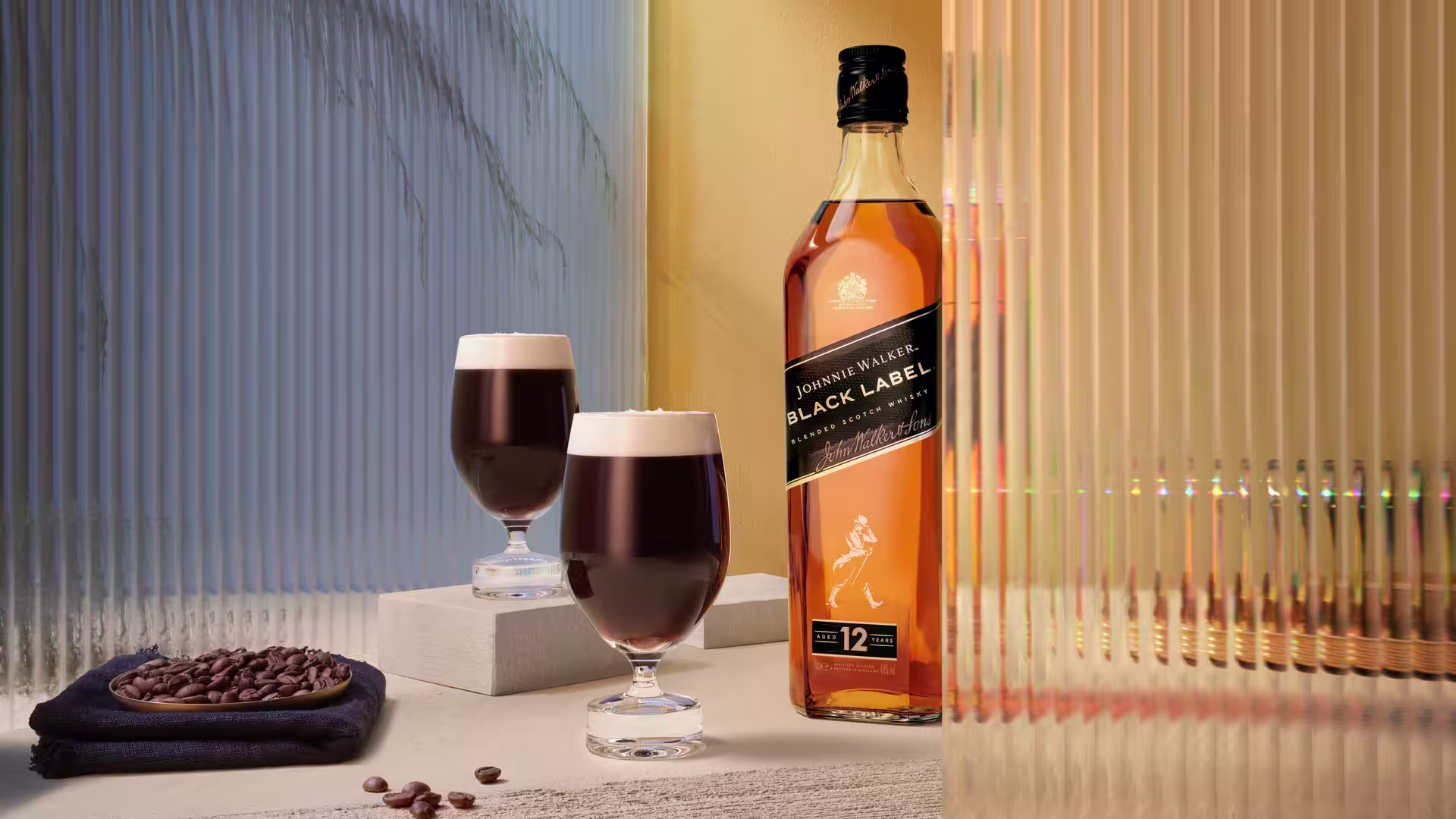 Una botella de whisky Johnnie Walker Black Label se encuentra en posición vertical junto a tres vasos llenos de un cóctel oscuro y cremoso, sobre un fondo de paneles de vidrio con rayas verticales.