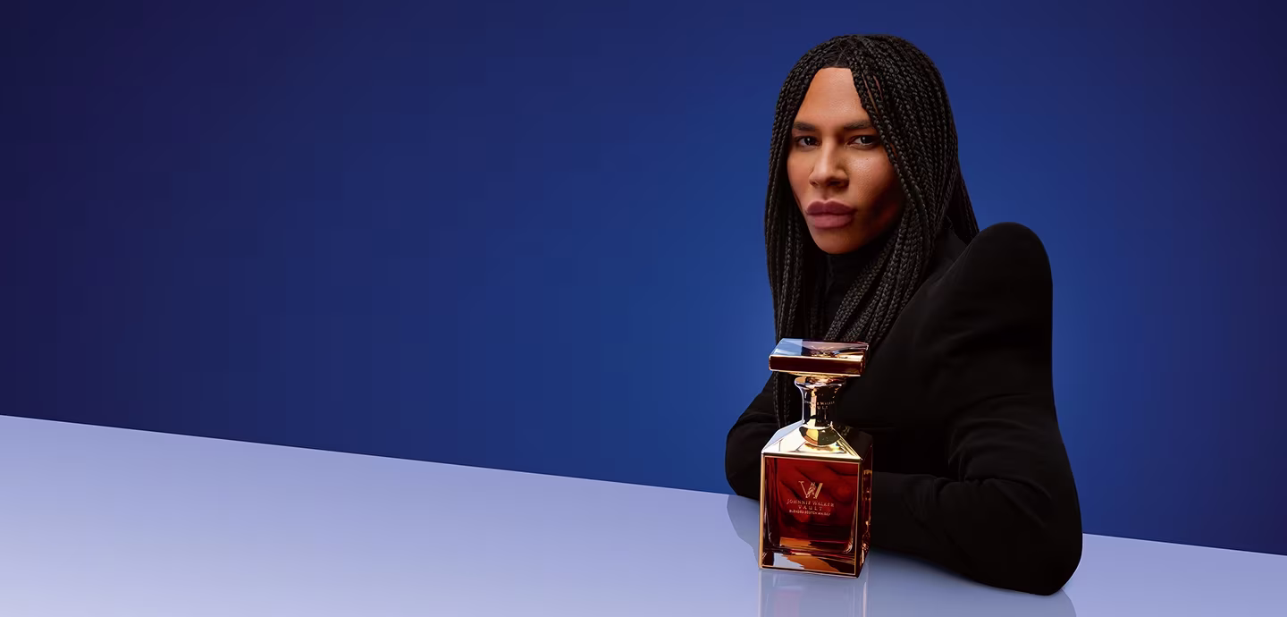 Person with long braids in black clothing poses with a square perfume bottle against a blue gradient background.