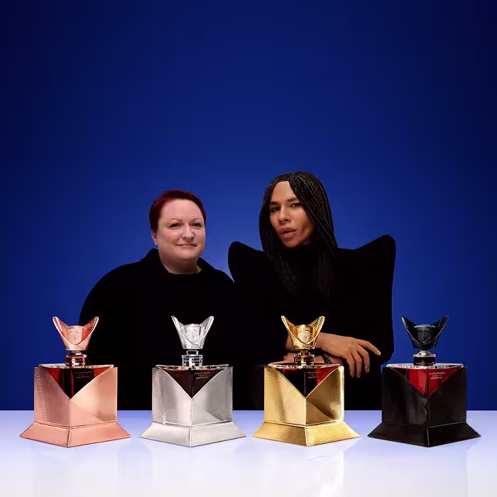 Hai người đứng trong một căn phòng sang trọng, sáng sủa với những kệ đựng chai nước hoa. Một người mặc vest đen và áo sơ mi trắng, người kia mặc váy đen. Những chiếc cốc thủy tinh được xếp trên một chiếc bàn gỗ trước mặt họ.