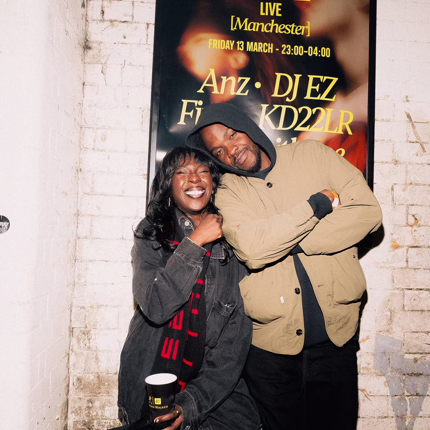 A man and woman pose in front of a poster for a live music event.