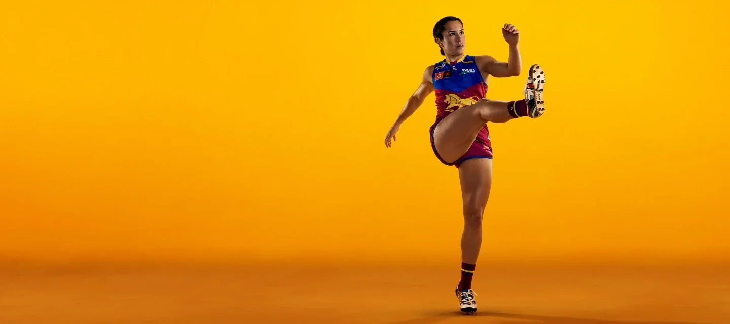 A person in a colourful sports uniform is in mid-air, kicking a ball against a bright yellow background. The uniform includes blue, red, and yellow colours, and the person wears sports shoes with cleats.