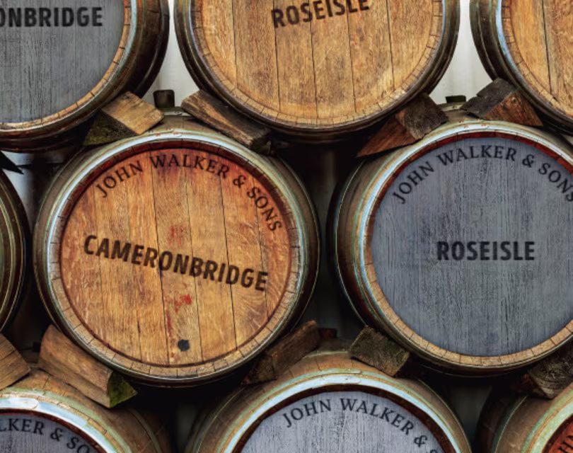 Close-up of stacked wooden barrels with the inscriptions Cameronbridge and Roseisle in black and brown text, surrounded by John Walker & Sons in a circular arrangement. The barrels vary in wood tone and texture.