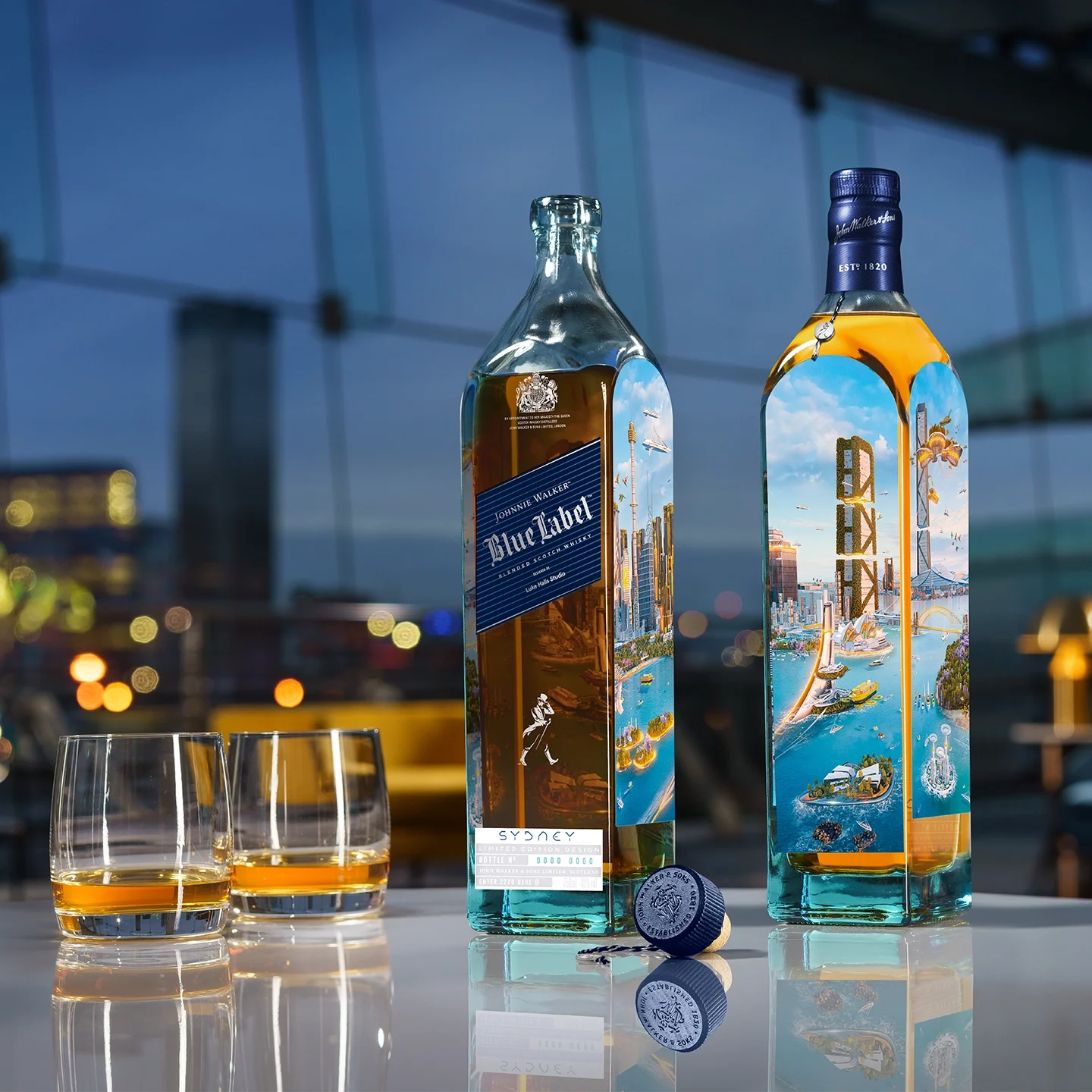 Two bottles of Johnnie Walker Blue Label whisky stand on a reflective table beside two glasses of whisky and a bottle cap, with a cityscape visible through large windows in the background.