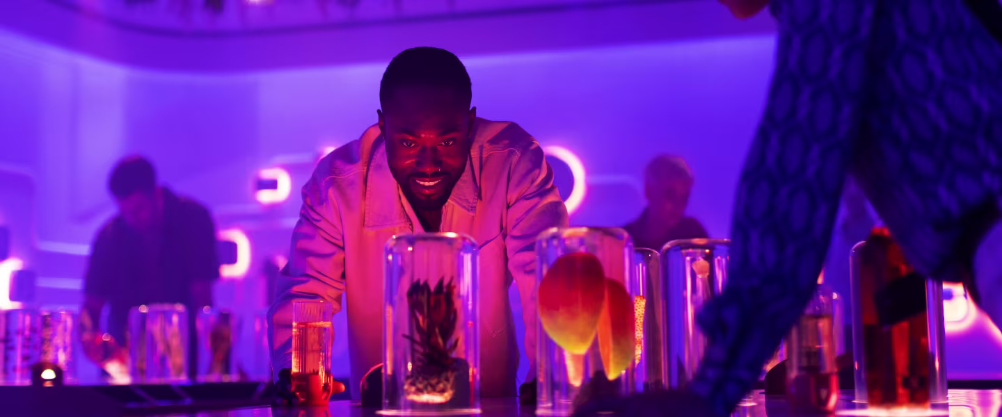 A man in a white shirt leans over a table with glass containers full of fruit under glowing purple and pink lights. Blurred figures and neon lights can be seen in the background, creating a vibrant, atmospheric backdrop.