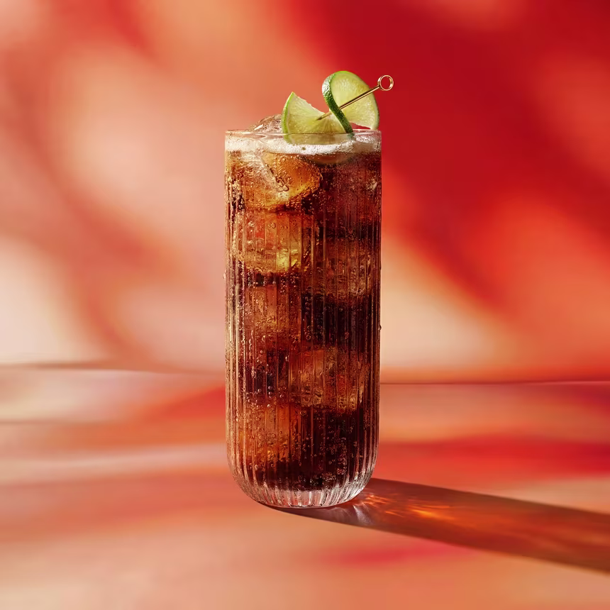 Vaso alto de cola con hielo, adornado con rodajas de lima, sobre un fondo difuminado de color naranja cálido y rojo.