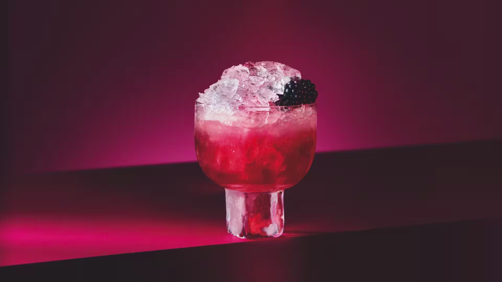 A glass filled with a red cocktail topped with crushed ice and garnished with a blackberry, set against a dark pink and red gradient background.