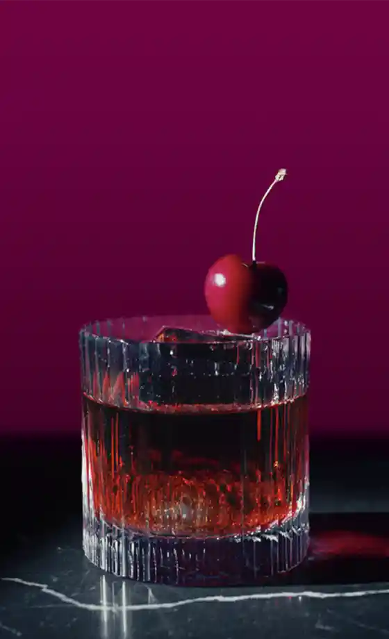 A glass of dark red liquid with ice stands on a marble surface against a dark pink background. A ripe cherry with a green stem is placed on the rim of the glass.