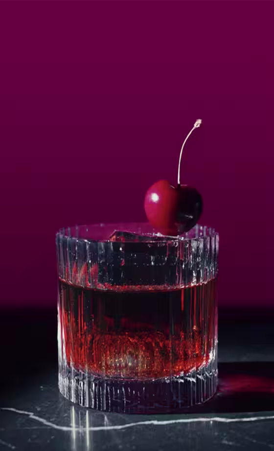 Un vaso de líquido rojo oscuro con hielo se encuentra sobre una superficie de mármol sobre un fondo rosa oscuro. En el borde de la copa se coloca una cereza madura con un tallo verde.