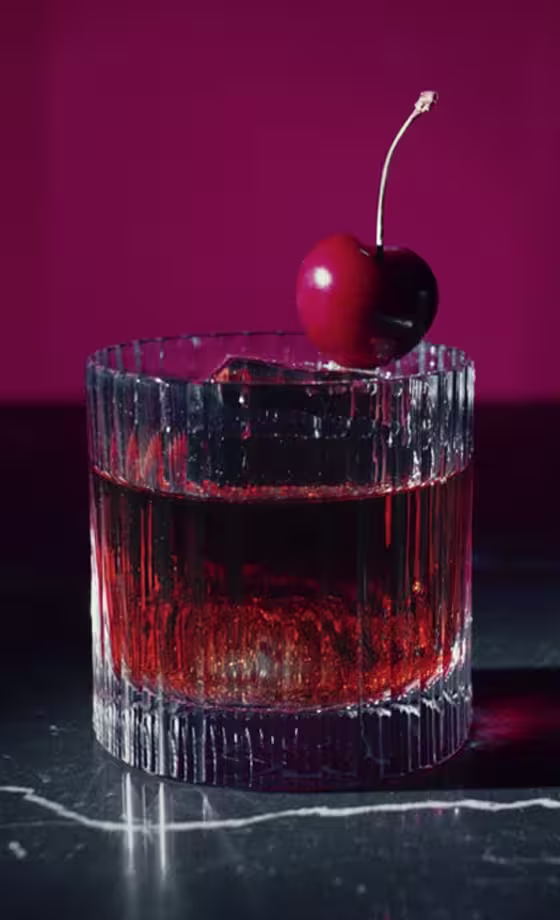 Un vaso con líquido rojo oscuro con un gran cubo de hielo y una cereza en el borde. El vaso tiene un patrón texturizado y está colocado sobre una superficie oscura y un fondo magenta que contrastan.