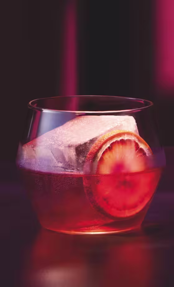 A glass of red cocktail with a large ice cube and an orange slice sits on a dark surface, with dramatic pink and purple lighting in the background.