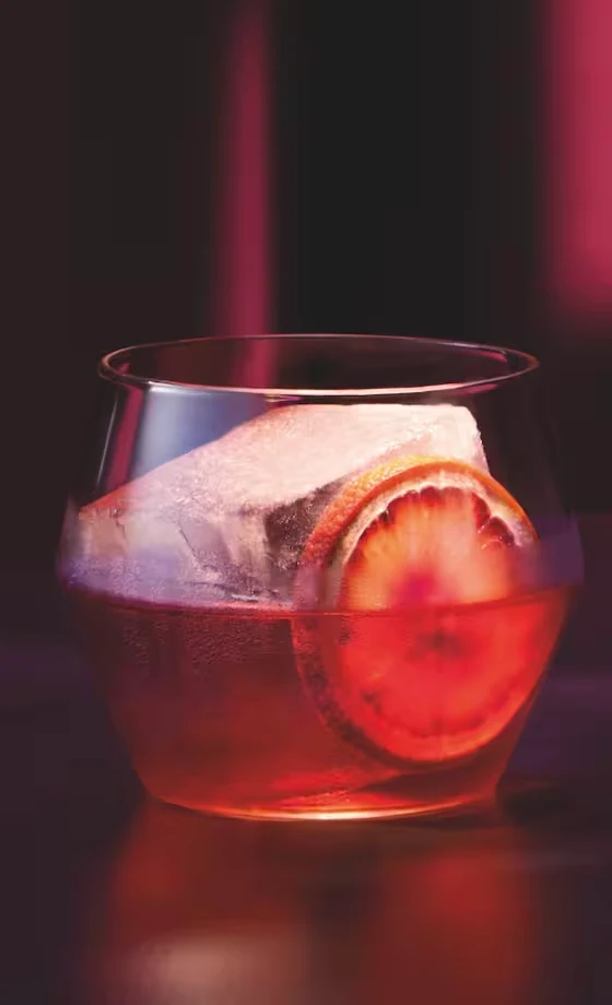 A glass of red cocktail with a large ice cube and an orange slice sits on a dark surface, with dramatic pink and purple lighting in the background.
