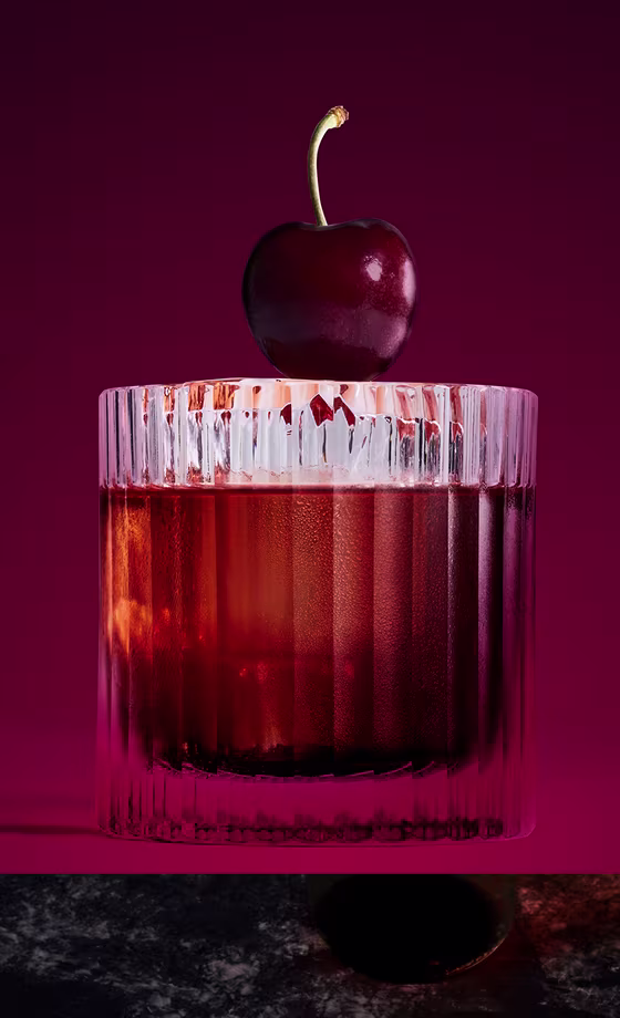A clear glass with a dark red cocktail sits on a marble surface. A single cherry is perched on the rim. The background is a deep purple, creating a dramatic atmosphere.