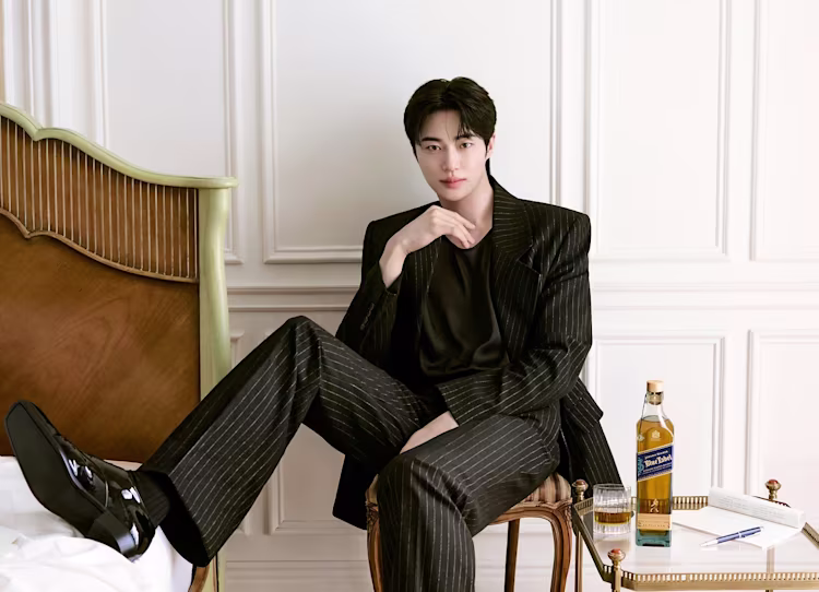 A man in a black pinstripe suit sits on a chair by a bed, with whisky and a glass on a small table beside him.