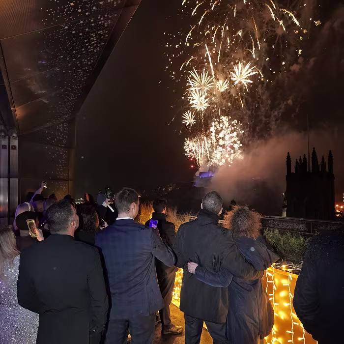 A group of people in formal wear watch fireworks outside at night, with string lights on a nearby railing.