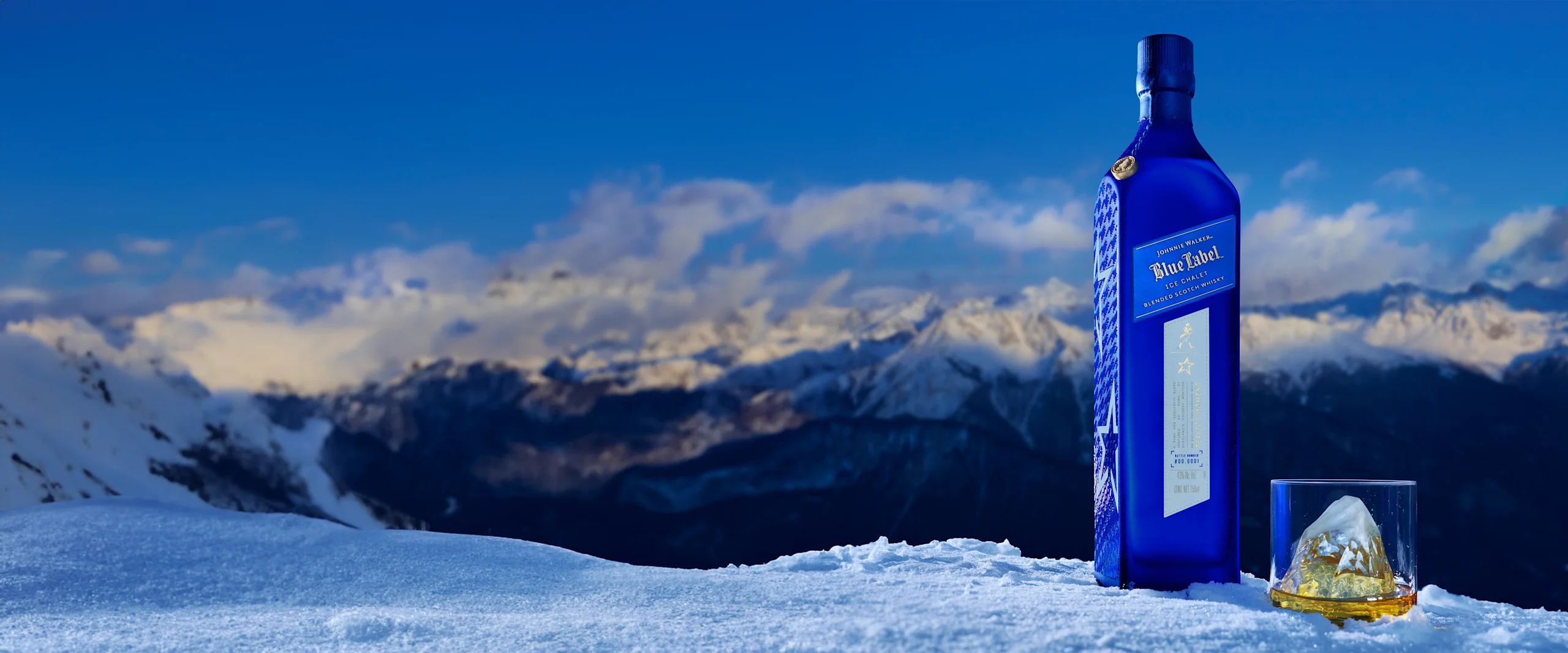 Johnnie Walker Blue Label Ice Chalet. A blue bottle of whiskey stands in snow with a glass filled with whiskey and a mountain-shaped ice cube. Snow-covered mountains and a bright blue sky are in the background.
