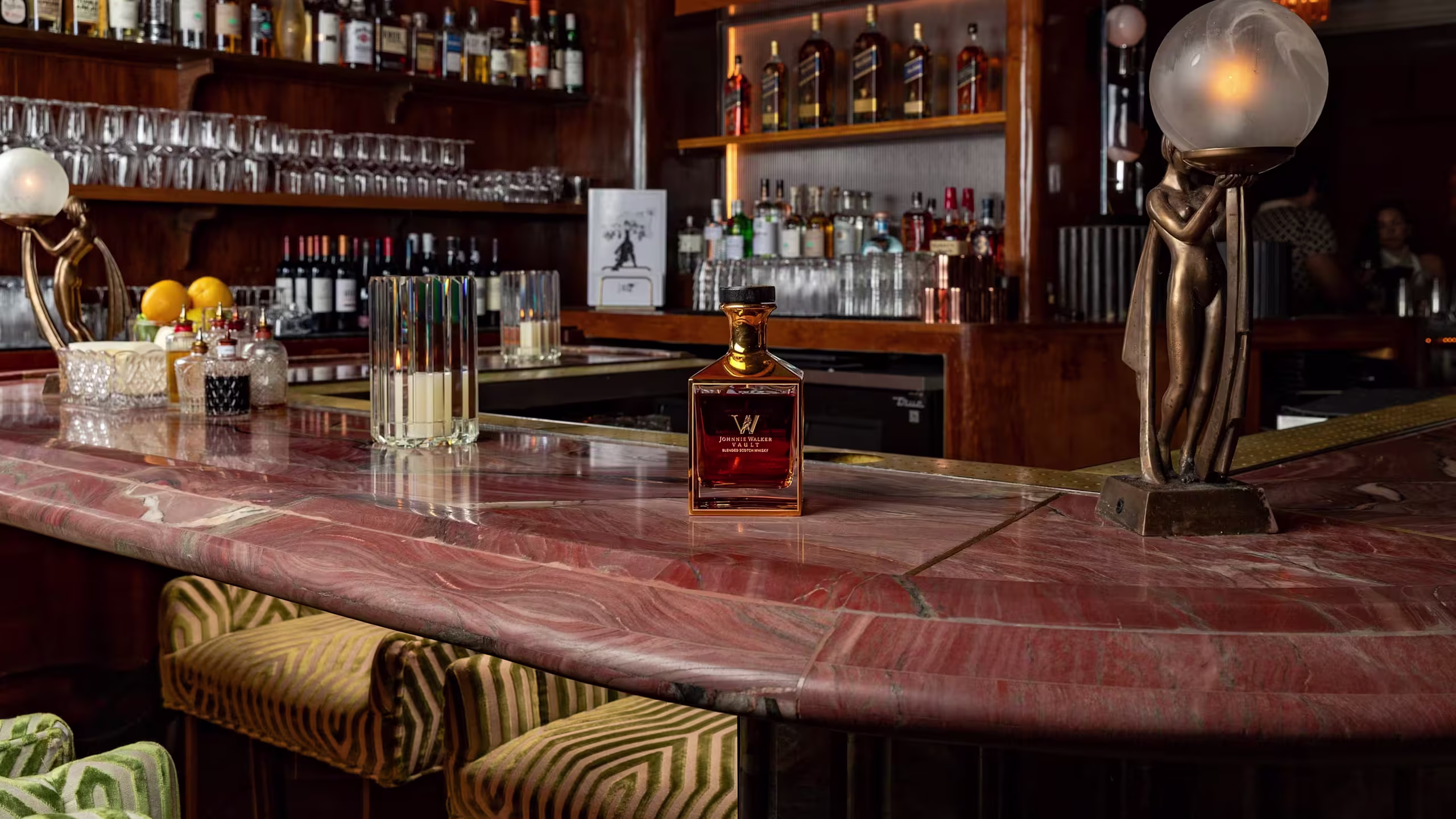 A bottle of whiskey sits on a marble bar counter in a dimly lit bar, with shelves of liquor, glassware, and art-deco style lamps in the background. Two patterned bar stools are partially visible at the counter.