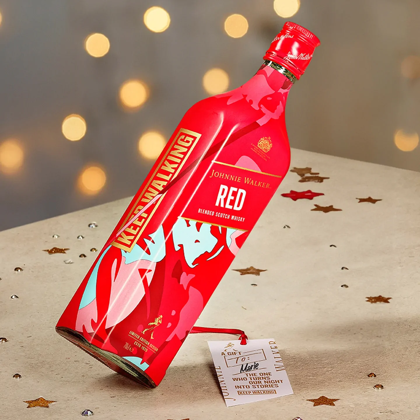 Бутилка уиски Johnnie Walker Red Label лежи на маса, украсена с конфети във формата на звезда. Бутилката е наситено червена с модерен флорален дизайн. Към гърлото на бутилката е прикрепен етикет с надписан текст.
