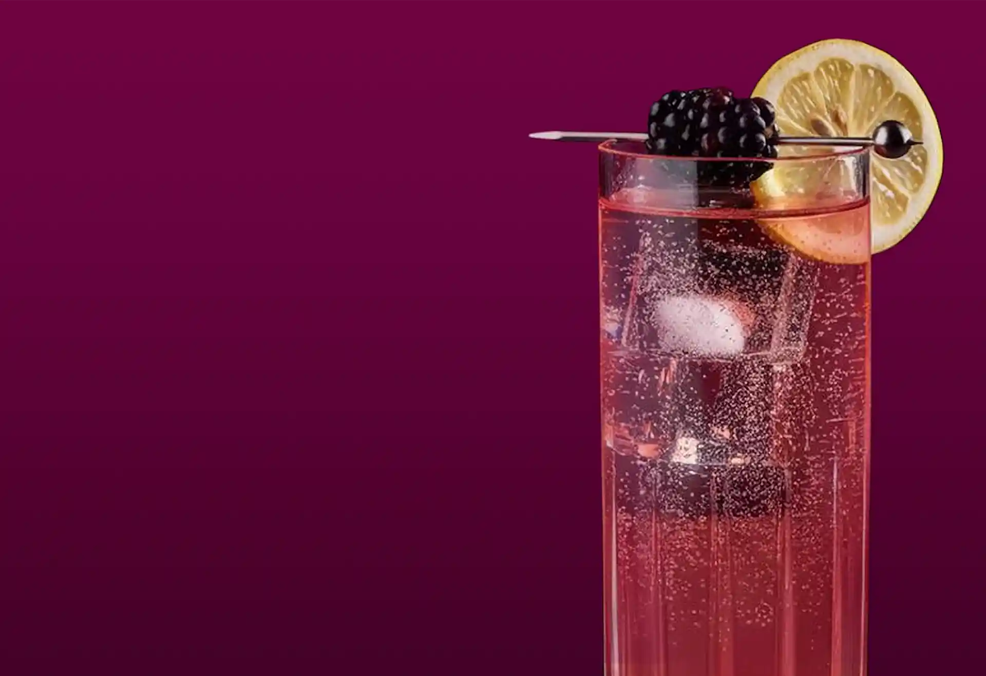 A sparkling pink cocktail in a tall glass with ice cubes, decorated with a lemon wedge and two blackberries, on a deep purple background.