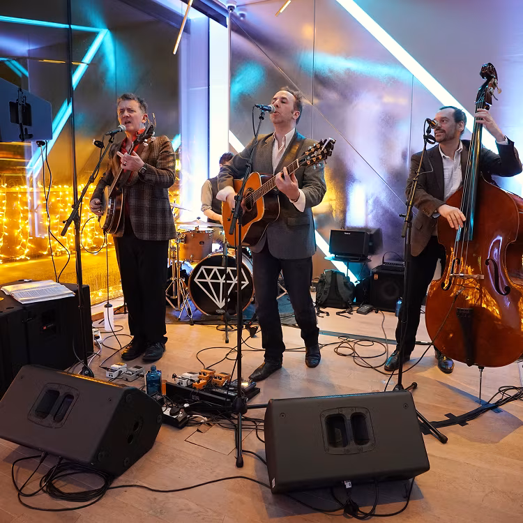 Three musicians perform on stage with guitar, double bass, and drums in a warmly lit indoor venue.