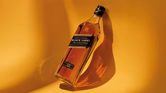 Uma garrafa de uísque escocês misturado Johnnie Walker Black Label está inclinada contra um fundo amarelo-dourado quente, projetando uma sombra clara.