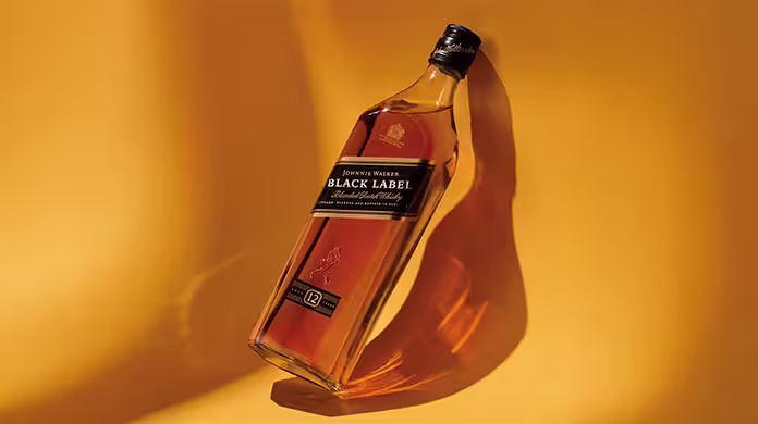 Бутилка шотландско уиски Johnnie Walker Black Label е наклонена на топъл, златистожълт фон, хвърляйки ясна сянка.