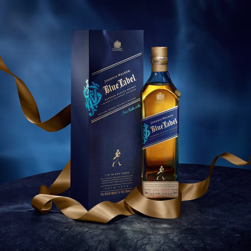 A bottle of Johnnie Walker Blue Label whisky stands beside its dark blue box packaging, both adorned with gold accents, on a marble surface with a gold ribbon elegantly draped around them.