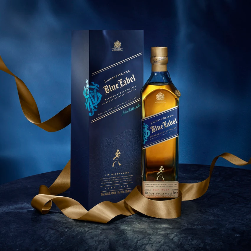 A bottle of Johnnie Walker Blue Label whisky stands beside its dark blue box packaging, both adorned with gold accents, on a marble surface with a gold ribbon elegantly draped around them.