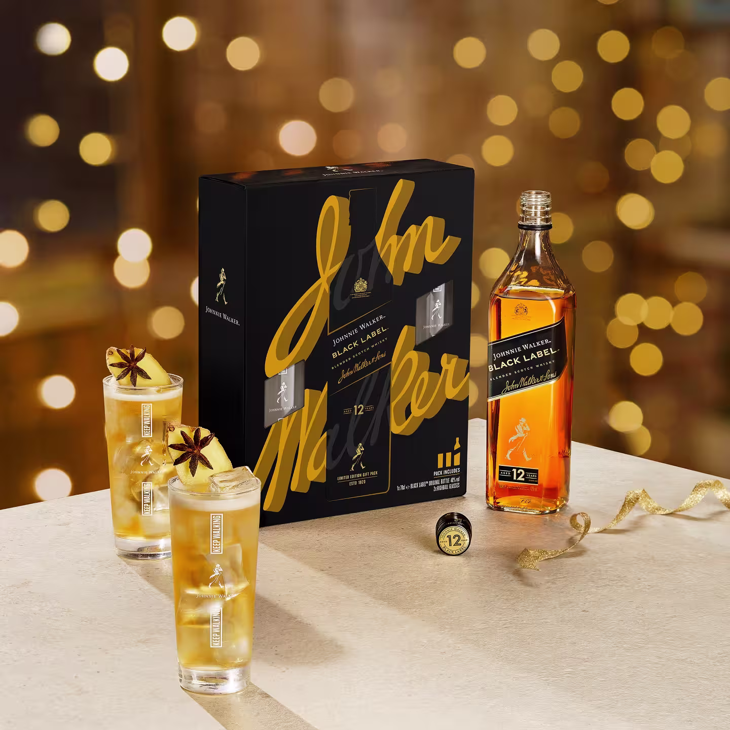 A bottle of Johnnie Walker Black Label whisky, two cocktails garnished with star anise, and a branded gift box are displayed on a table with a festive, blurred lights background.