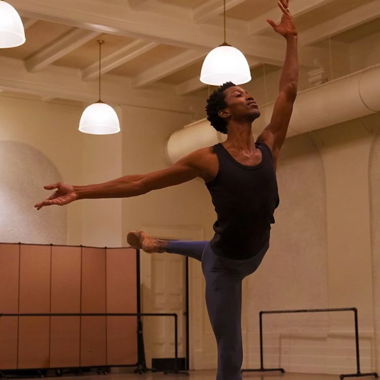 A ballet dancer in a black tank top and blue leggings performs an elegant arabesque in a spacious dance studio with high ceilings and hanging lights. The room features a folding screen and barre in the background.