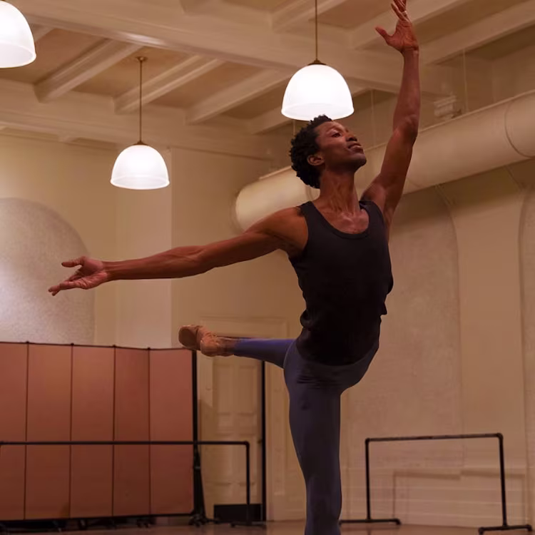 Una bailarina de ballet con una camiseta negra y medias azules realiza un arabesco en un espacioso estudio con techos altos y luces colgantes.