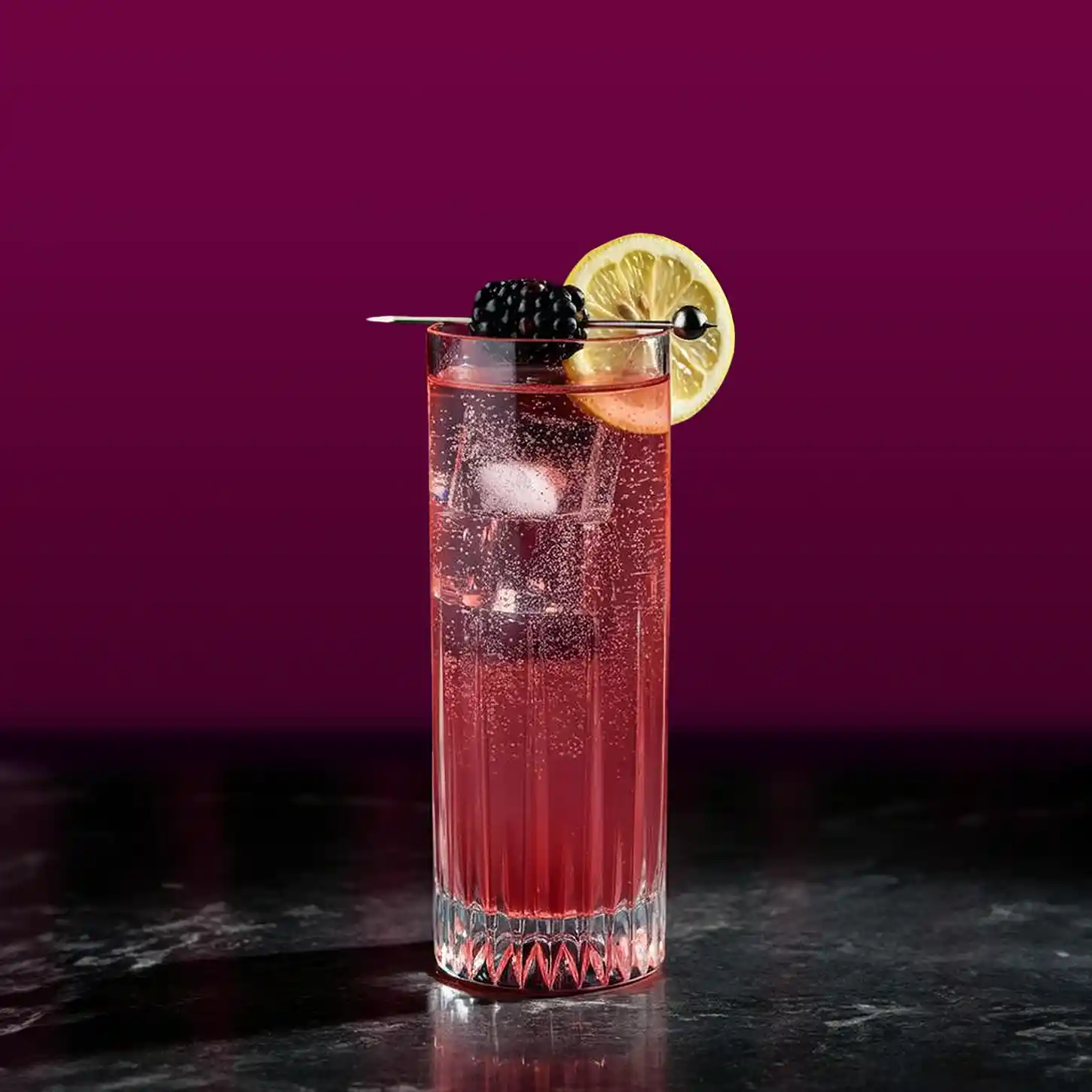 A tall, clear glass filled with a fizzy pink cocktail garnished with a lemon slice and a blackberry on a skewer. The drink is set against a deep purple background on a dark surface.